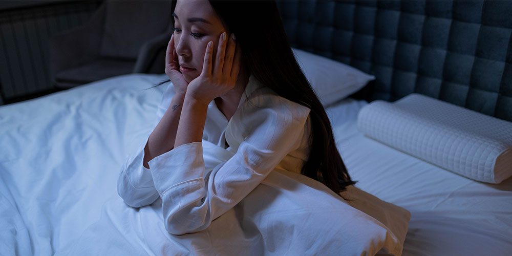 woman in white long sleeve shirt sitting on a bed having difficulty sleeping
